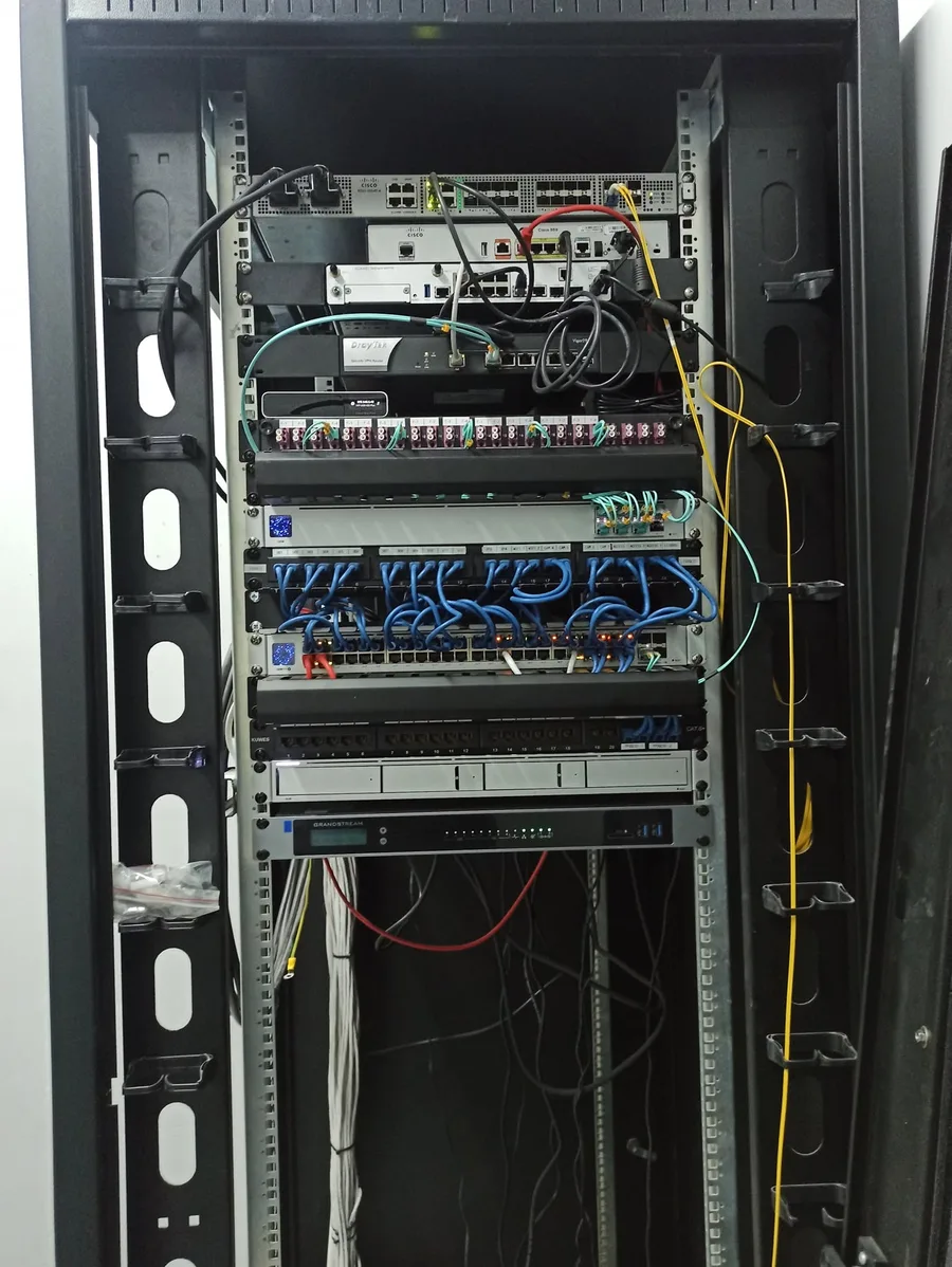 Ανοιχτό rack με router, switch, κατανεμητή οπτικών ινών, patch panel Cat6 με μπλε καλώδια και τηλεφωνικό κέντρο VoIP.